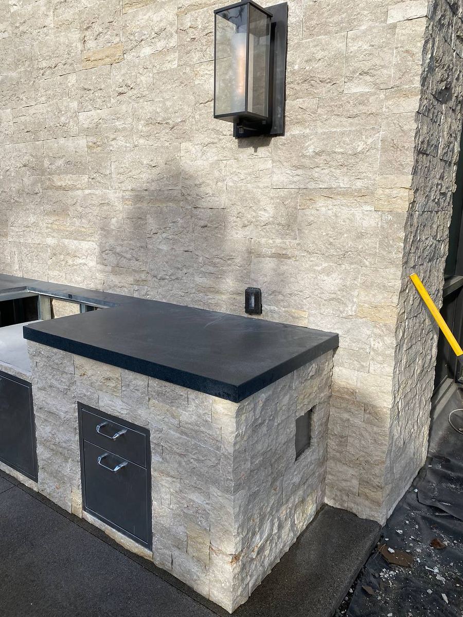 Outdoor kitchen island clad in split-face stone with a black stone countertop against a matching stone wall